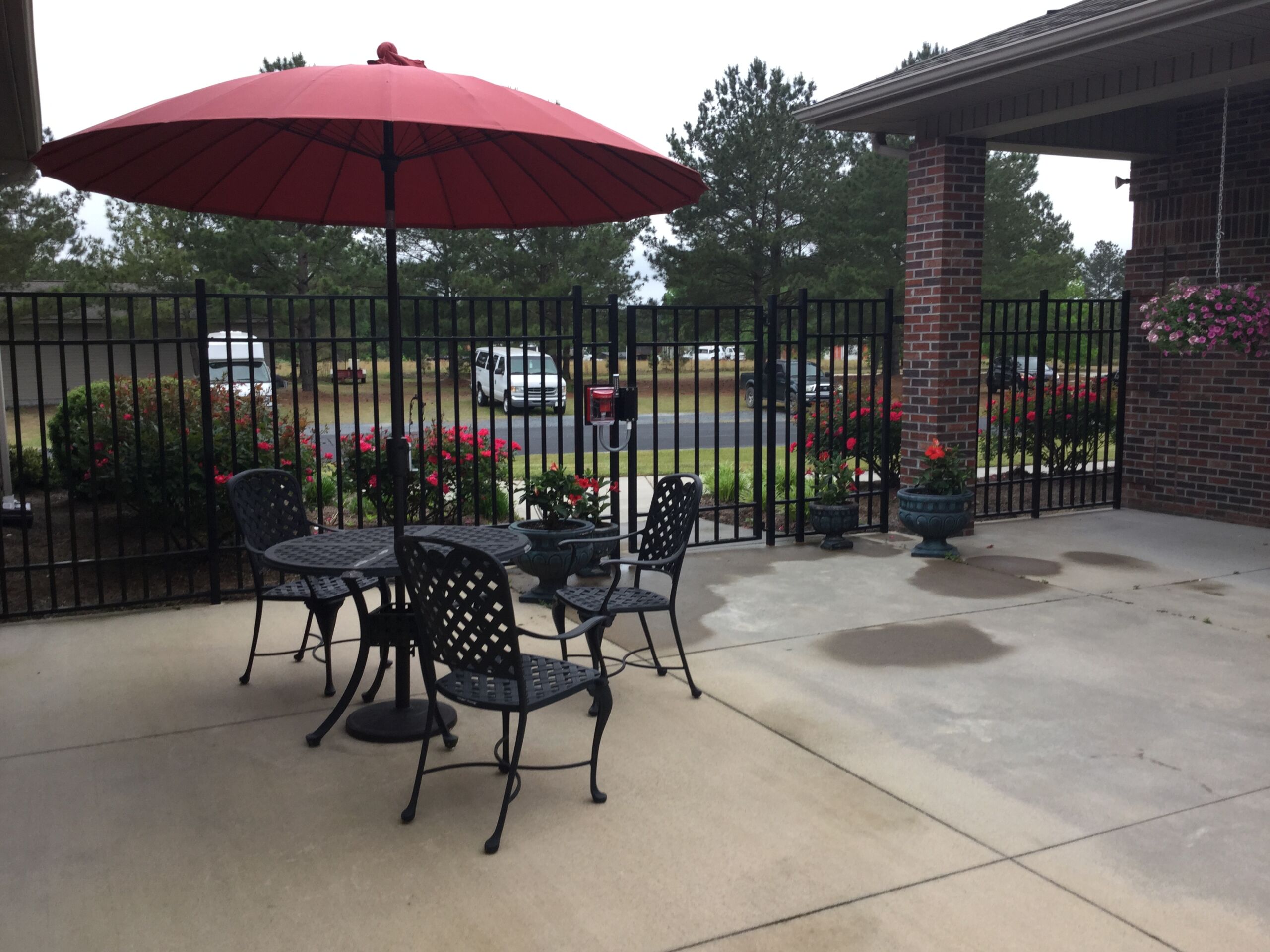 Outdoor seating area featuring a round table and chairs under a large red umbrella, surrounded by flower beds and a gated fence.