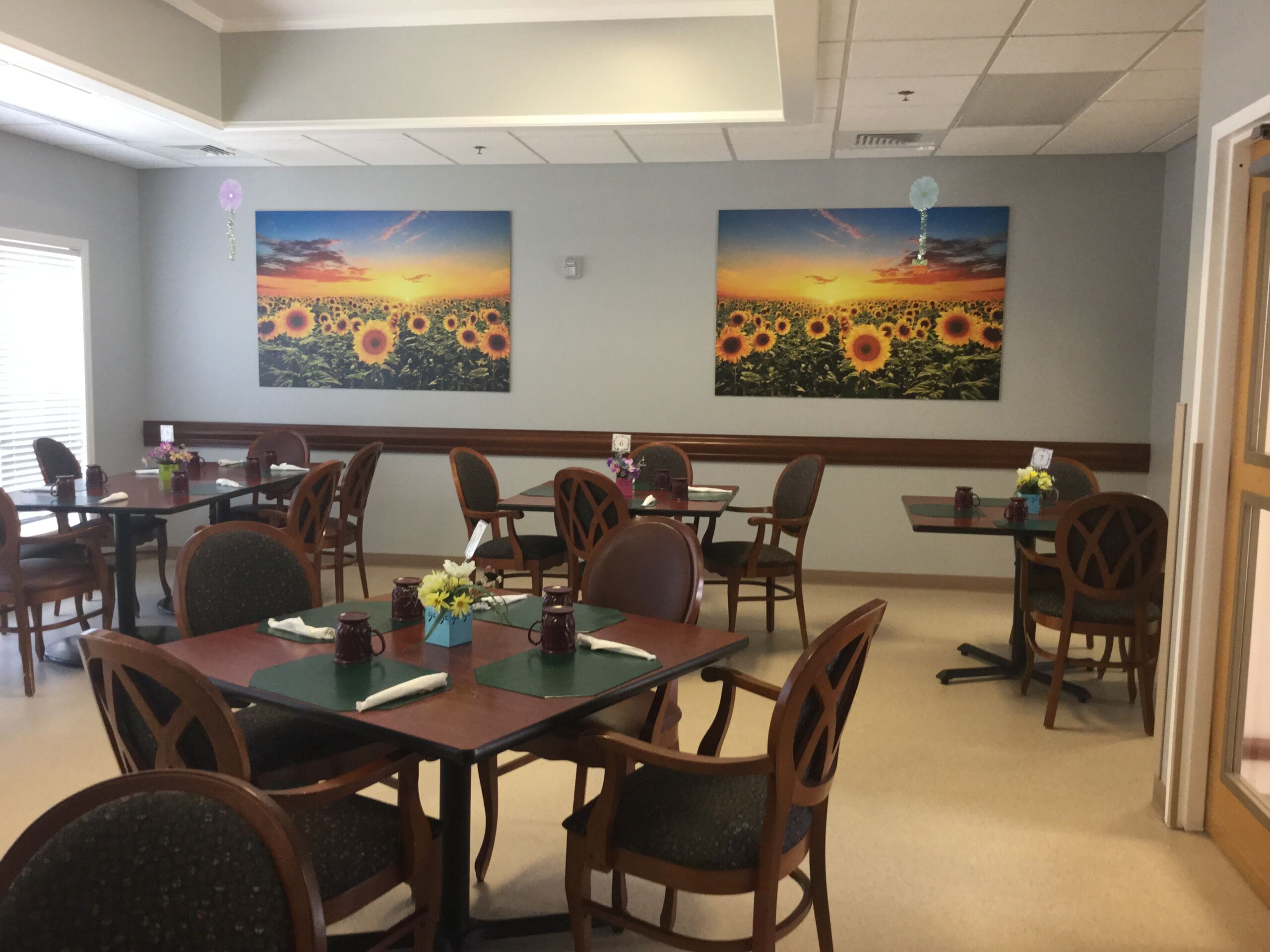 Bright dining area featuring tables set with flowers, green placemats, and framed sunflower artwork on the walls, creating a welcoming atmosphere.