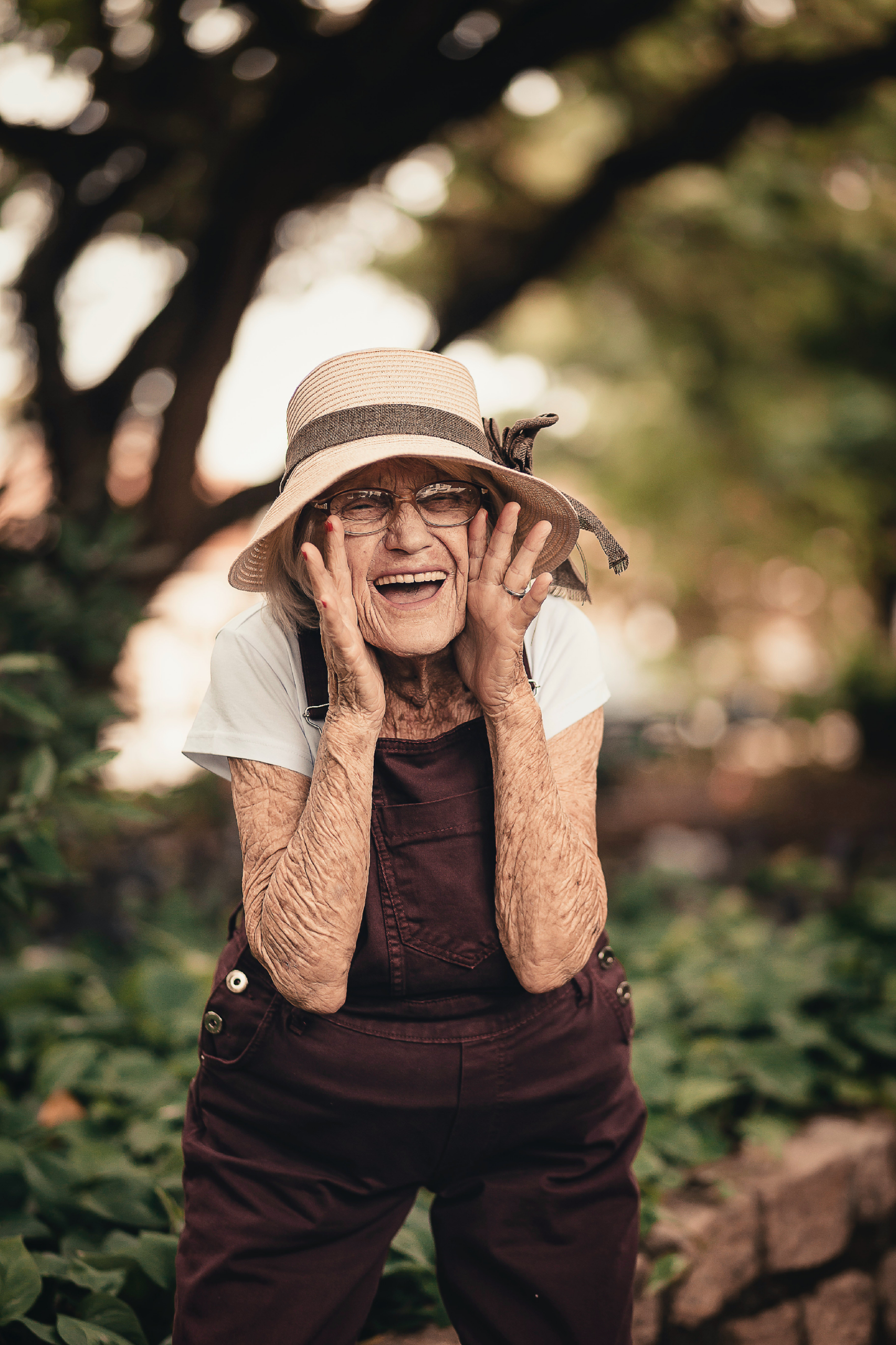 An elderly woman in a wide-brimmed hat joyfully poses outdoors, smiling and gesturing playfully among greenery. Her expression conveys happiness and warmth.