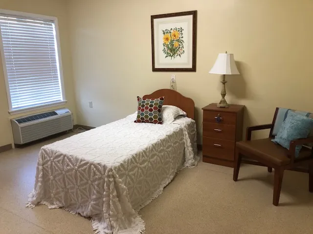 Cozy bedroom featuring a single bed with a decorative throw and pillow, a nightstand with a lamp, and a chair. Natural light streams in through a window.