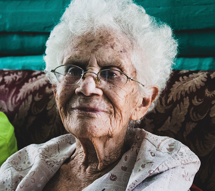 Elderly woman with curly white hair and glasses smiles warmly while seated on a couch. Her expression conveys wisdom and serenity.